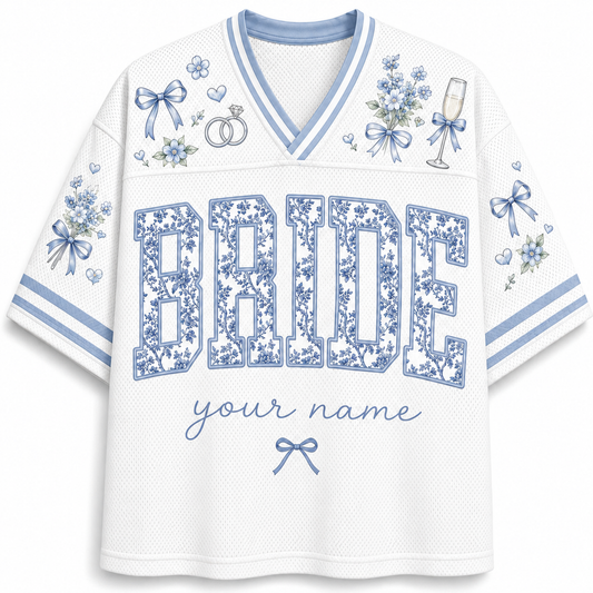 White mesh V-neck jersey-style shirt with blue floral print details, including bows, flowers, rings, a champagne glass, and large 'BRIDE' lettering with placeholder text 'your name' on the front.