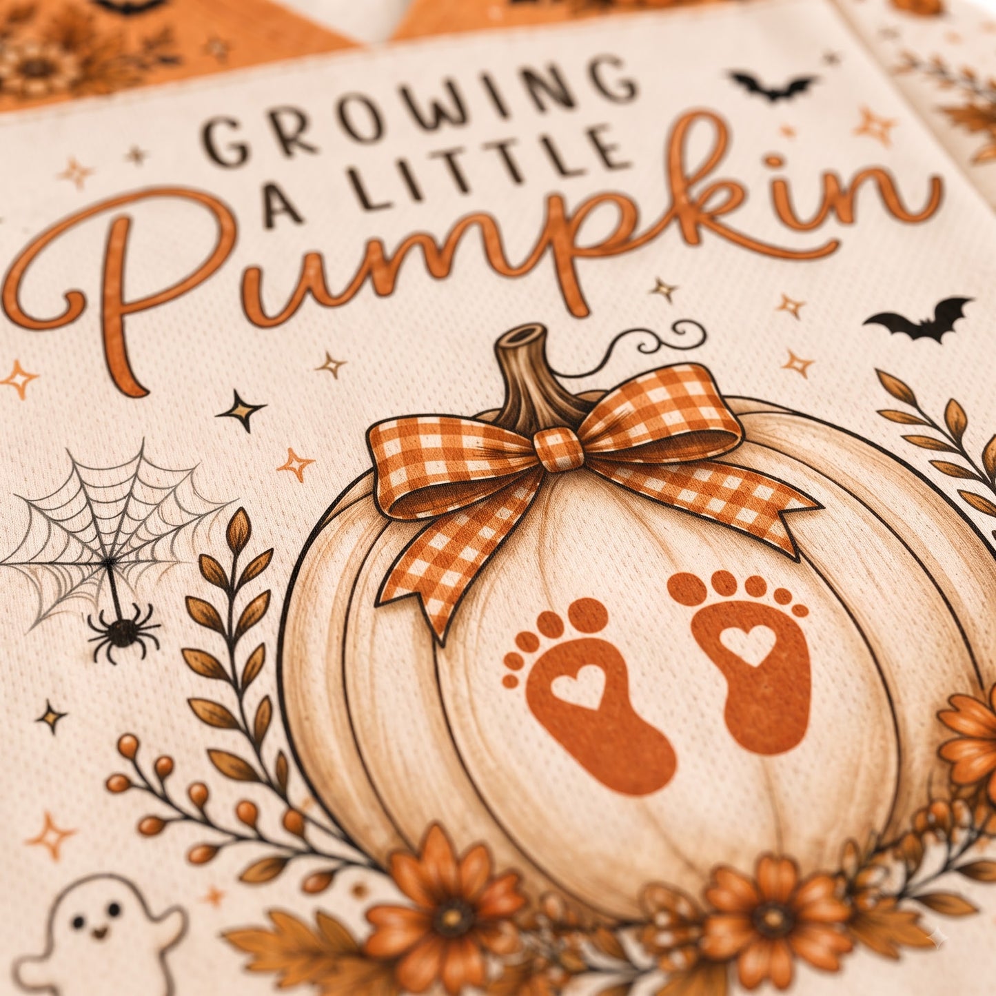 Close-up of a printed pumpkin-themed design with the phrase “Growing a Little Pumpkin” in brown lettering, a large cream pumpkin illustration with an orange gingham bow, orange baby footprints, autumn leaves, flowers, spiders, bats, and a ghost.