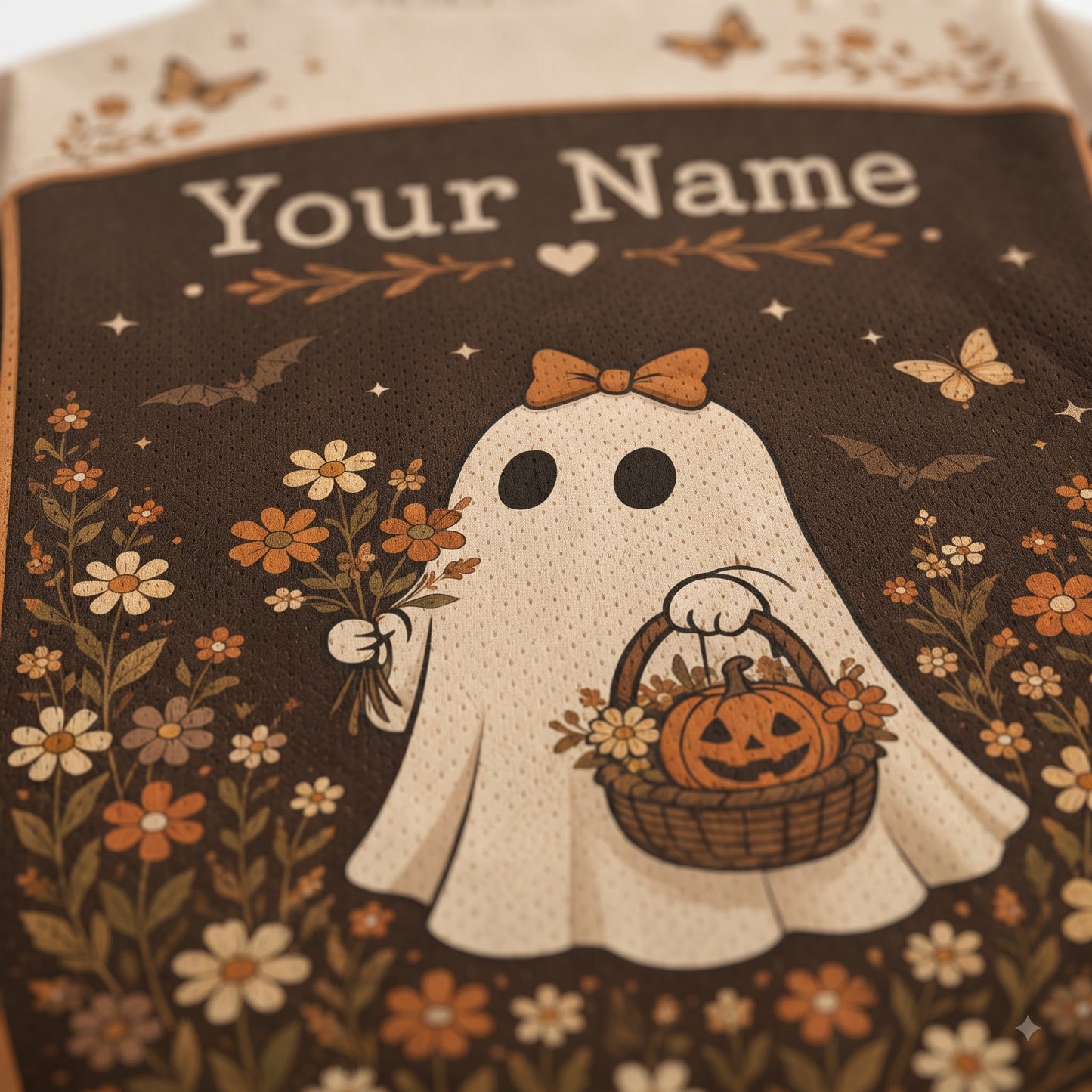Close-up of a personalized Halloween-themed fabric item, likely a bag or pouch, featuring a cream-colored ghost graphic with the text 'Your Name' at the top. The design includes a pumpkin basket, flowers, butterflies, and bats on a dark brown background.