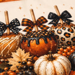 A close-up styled arrangement of autumn pumpkins and decorative bows on a warm beige background. The pumpkins include orange drizzled candy corn, a cream pumpkin with black speckles, a leopard-print pumpkin, and others with black bows, autumn leaves, and orange berries.