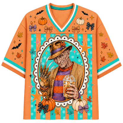 An orange short-sleeve V-neck jersey-style shirt with a Halloween-themed graphic print. The design features a central oval illustration of a horror character in a hat holding a drink, surrounded by pumpkins, bats, stars, leaves, bows, and teal and white striped accents.