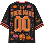 A black mesh short-sleeve jersey shown from the back, featuring customizable orange and pink text reading 'YOUR NAME' above large '00' numbers. The shirt is decorated with autumn-themed graphics including pumpkins, bows, leaves, stars, a witch hat, and a cauldron, with orange, pink, and black striped sleeve cuffs.