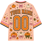 A peach-colored customizable baseball-style jersey shown from the back, featuring orange and black 'YOUR NAME' lettering above large '00' numbers. The design includes orange and white pumpkins, pink bow motifs, autumn leaves, and small stars, with striped collar and sleeve cuffs.