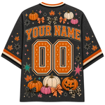 A black mesh short-sleeve jersey-style shirt shown from the back, decorated with orange and white pumpkins, pink pumpkins, plaid bows, stars, and the text 'YOUR NAME' above large '00' numbers. The shirt has orange, white, and black striped sleeve cuffs and a white collar trim.