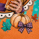Close-up of a Halloween-themed printed surface featuring a large orange pumpkin with a purple gingham bow, autumn maple leaves, a skeleton hand holding a mug, and an orange textured background.