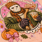 A close-up of a Halloween-themed illustrated design featuring a masked figure in an olive green jacket and orange plaid scarf, holding a jack-o'-lantern cup, with pumpkins, leaves, stars, and autumn decorations on a pink patterned background.