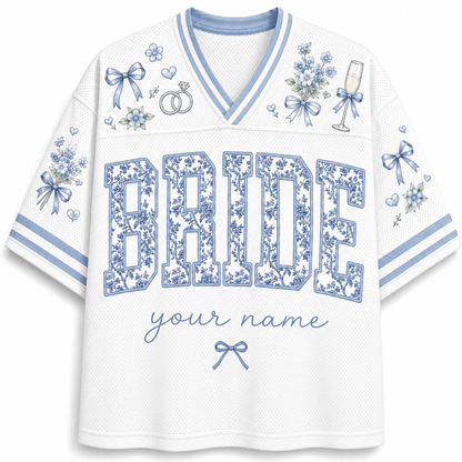 White mesh V-neck jersey-style shirt with blue floral print details, including bows, flowers, rings, a champagne glass, and large 'BRIDE' lettering with placeholder text 'your name' on the front.