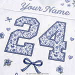 Close-up of a white personalized sports jersey fabric with blue floral patterned number 24, blue script 'Your Name' at the top, small blue flowers and hearts, and a blue bow graphic.