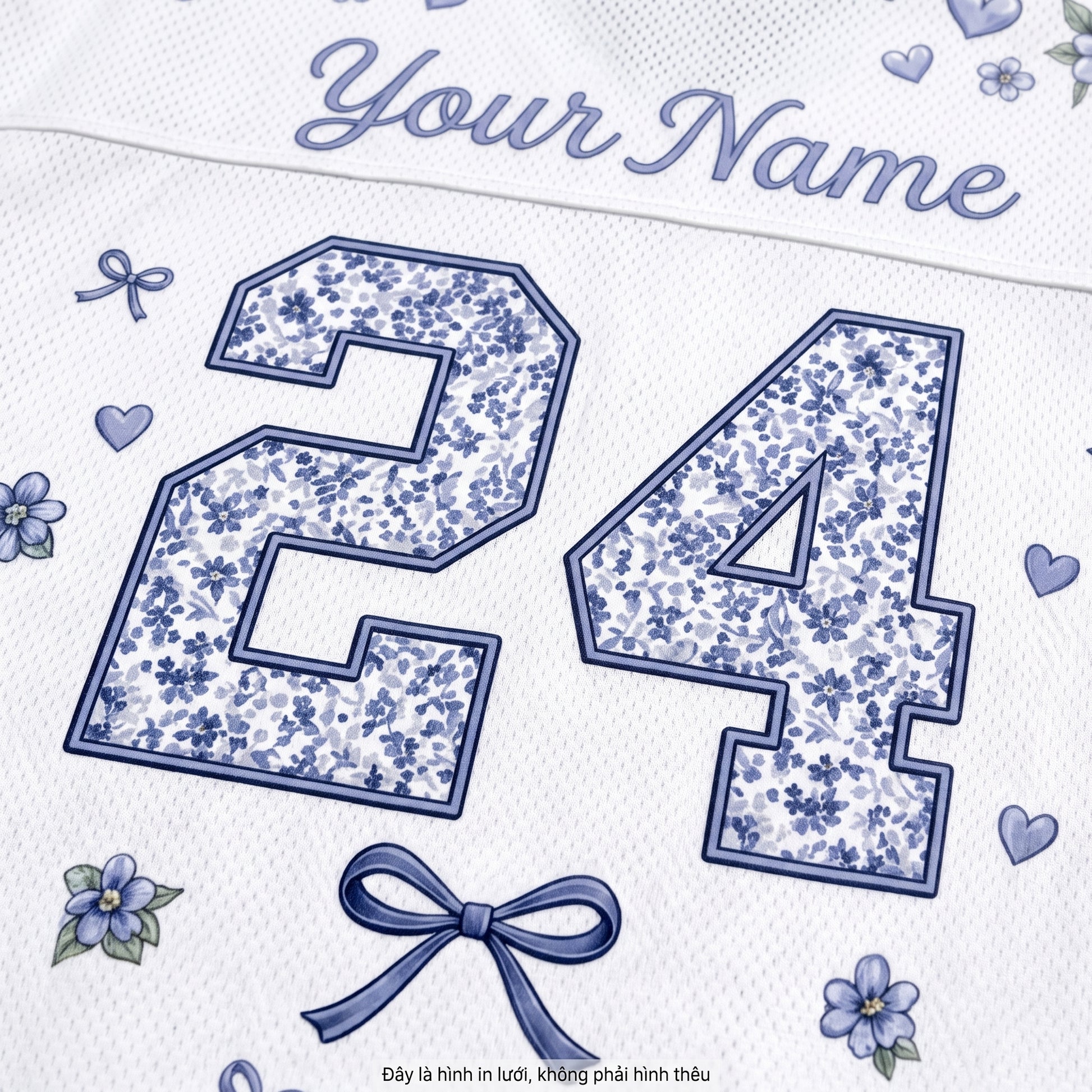 Close-up of a white personalized sports jersey fabric with blue floral patterned number 24, blue script 'Your Name' at the top, small blue flowers and hearts, and a blue bow graphic.