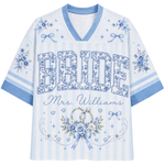 A women's short-sleeve V-neck t-shirt in light blue and white, featuring 'BRIDE Mrs. Williams' printed in floral and script fonts, decorated with wedding rings, flowers, and ribbons.