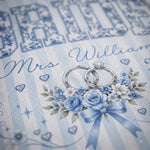 A close-up of a printed wedding invitation or card design featuring the text 'Mrs. William' in cursive, large decorative letters at the top, two interlocking wedding rings, and a bouquet of blue and white flowers with a blue ribbon on a pale blue striped background.