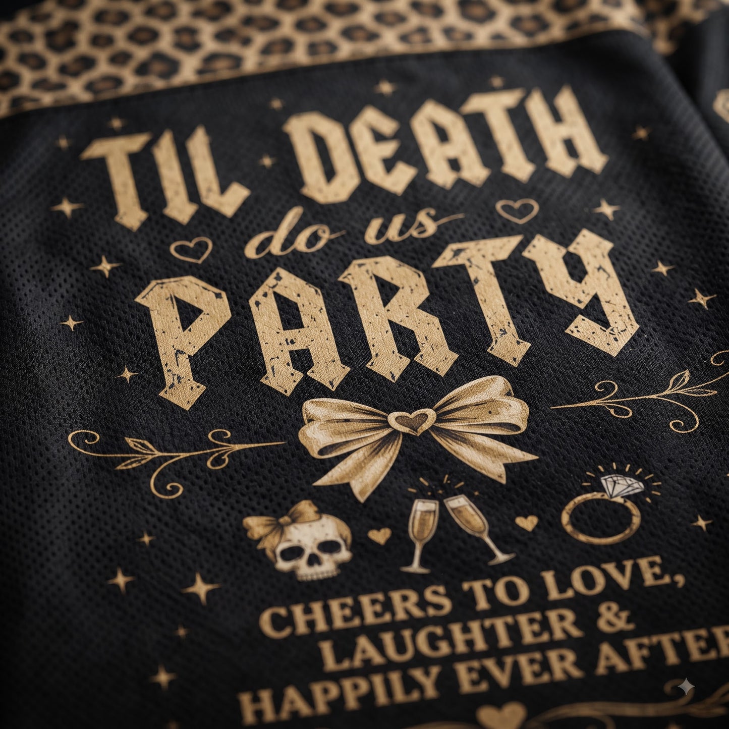 Close-up of a black mesh fabric item printed with distressed gold gothic text reading 'TIL DEATH do us PARTY' and additional celebratory graphics including a bow, skulls, champagne glasses, rings, and stars, with leopard-print fabric visible at the top edge.