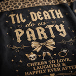 Close-up of a black mesh fabric item printed with distressed gold gothic text reading 'TIL DEATH do us PARTY' and additional celebratory graphics including a bow, skulls, champagne glasses, rings, and stars, with leopard-print fabric visible at the top edge.