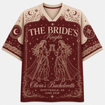 A short-sleeve V-neck t-shirt with a bachelorette graphic design. The shirt features a deep burgundy red body with cream-colored text reading 'THE BRIDE's Knights' and 'Olivia's Bachelorette', along with line-art illustrations of two knights holding cups under a crescent moon and stars.