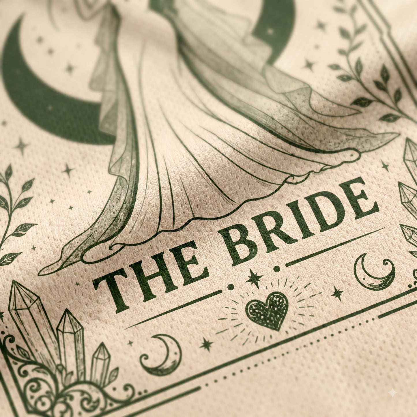 A close-up of a beige fabric napkin or paper towel printed with an elegant gothic-style illustration of a bride in a flowing gown, surrounded by crescent moons, stars, crystals, and the words 'THE BRIDE' in dark green lettering.