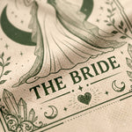 A close-up of a beige fabric napkin or paper towel printed with an elegant gothic-style illustration of a bride in a flowing gown, surrounded by crescent moons, stars, crystals, and the words 'THE BRIDE' in dark green lettering.