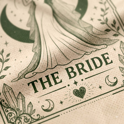 A close-up of a beige fabric napkin or paper towel printed with an elegant gothic-style illustration of a bride in a flowing gown, surrounded by crescent moons, stars, crystals, and the words 'THE BRIDE' in dark green lettering.