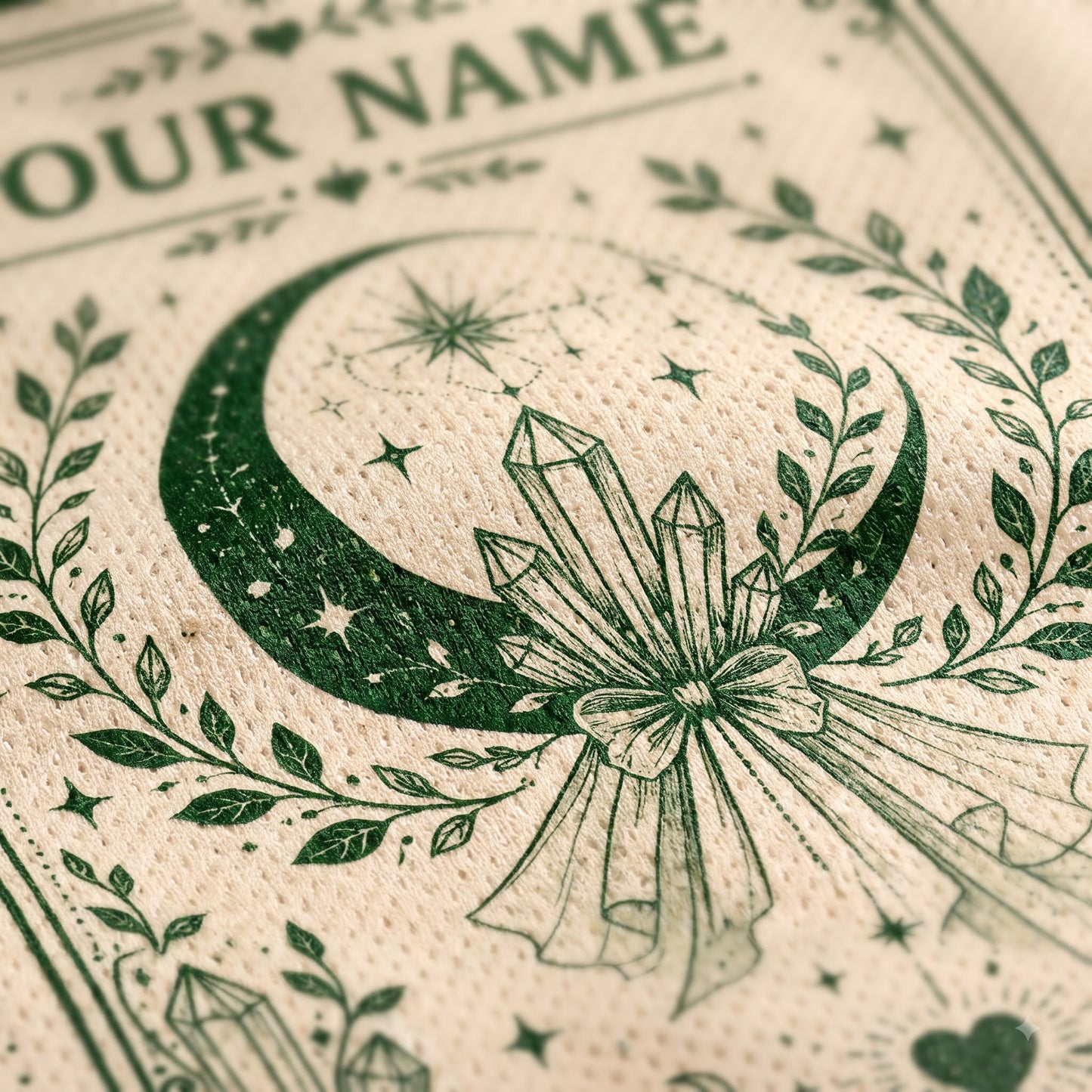 Close-up of a cream-colored textured paper napkin printed with a dark green occult-style design featuring a crescent moon, stars, crystals, and a bow, with partial text reading 'OUR NAME' at the top.