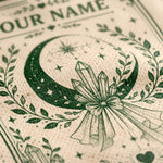 Close-up of a cream-colored textured paper napkin printed with a dark green occult-style design featuring a crescent moon, stars, crystals, and a bow, with partial text reading 'OUR NAME' at the top.
