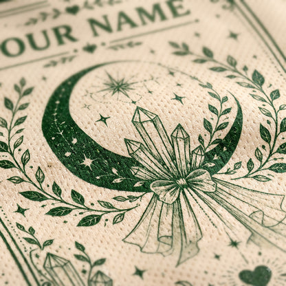 Close-up of a cream-colored textured paper napkin printed with a dark green occult-style design featuring a crescent moon, stars, crystals, and a bow, with partial text reading 'OUR NAME' at the top.