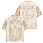 Front and back views of a custom short-sleeve V-neck jersey in cream and gold, featuring celestial designs including crescent moons, stars, and floral borders. The front displays an illustration of a bride in a forest with the text 'THE BRIDE', and the back shows 'YOUR NAME' in a decorative banner.
