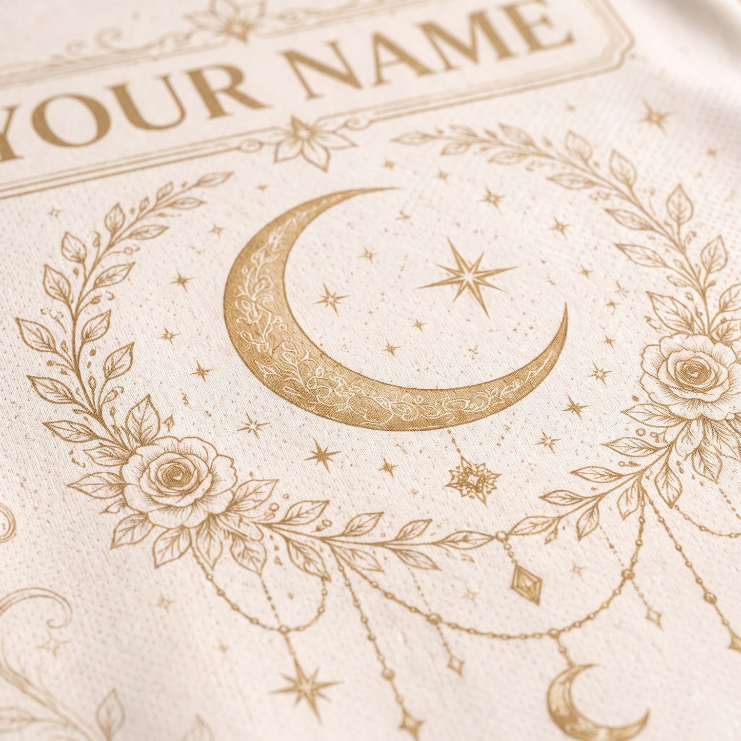 Close-up of a cream-colored textile featuring a gold-toned crescent moon, stars, floral wreath, and decorative text that reads 'YOUR NAME'.