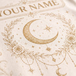 Close-up of a cream-colored textile featuring a gold-toned crescent moon, stars, floral wreath, and decorative text that reads 'YOUR NAME'.