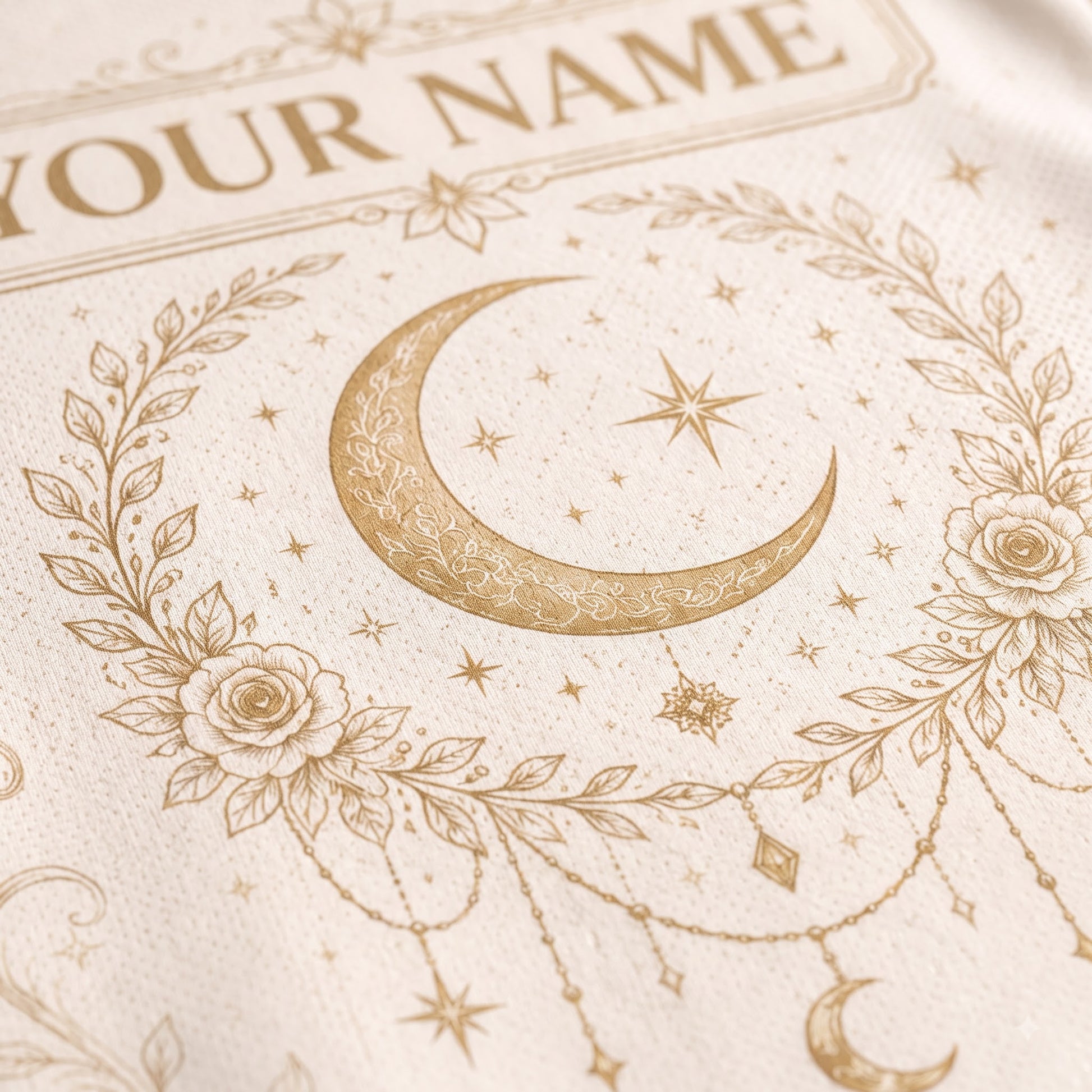 Close-up of a cream-colored textile featuring a gold-toned crescent moon, stars, floral wreath, and decorative text that reads 'YOUR NAME'.