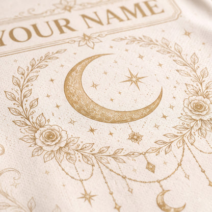 Close-up of a cream-colored textile featuring a gold-toned crescent moon, stars, floral wreath, and decorative text that reads 'YOUR NAME'.