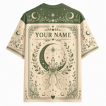 A short-sleeve t-shirt with a celestial and occult-themed graphic print. The shirt features a dark olive green yoke and sleeves with moon and star illustrations, and a large cream-colored central panel with 'YOUR NAME' printed in gothic lettering above a large crescent moon and crystal design.