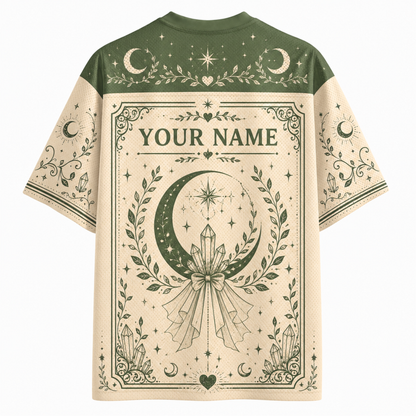 A short-sleeve t-shirt with a celestial and occult-themed graphic print. The shirt features a dark olive green yoke and sleeves with moon and star illustrations, and a large cream-colored central panel with 'YOUR NAME' printed in gothic lettering above a large crescent moon and crystal design.