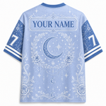 A light blue short-sleeve athletic-style shirt shown from the back, featuring a white celestial print with a crescent moon, stars, floral wreath, and the text 'YOUR NAME' across the upper back. The sleeves are blue with white number 7 graphics and striped cuffs.