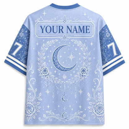 A light blue short-sleeve athletic-style shirt shown from the back, featuring a white celestial print with a crescent moon, stars, floral wreath, and the text 'YOUR NAME' across the upper back. The sleeves are blue with white number 7 graphics and striped cuffs.