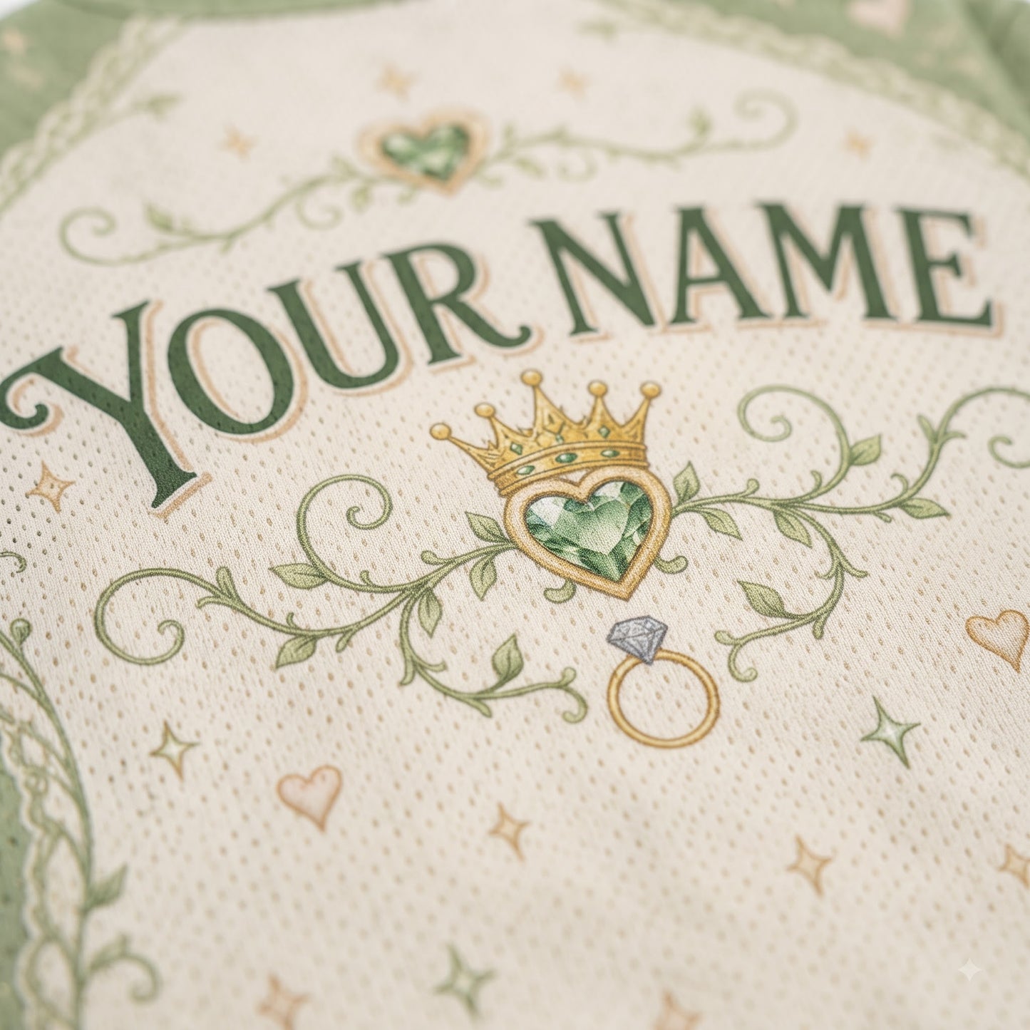 Close-up of a personalized cream-colored fabric item, likely a baby blanket or swaddle, featuring embroidered green text reading 'YOUR NAME', a green heart-shaped gem crown, and a gold ring motif.