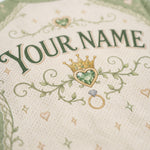 Close-up of a personalized cream-colored fabric item, likely a baby blanket or swaddle, featuring embroidered green text reading 'YOUR NAME', a green heart-shaped gem crown, and a gold ring motif.