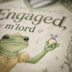 Close-up of a cream-colored mesh garment featuring a printed illustration of a green frog wearing a gold crown and holding a diamond ring, with the text 'Engaged, m'lord' in dark green script.