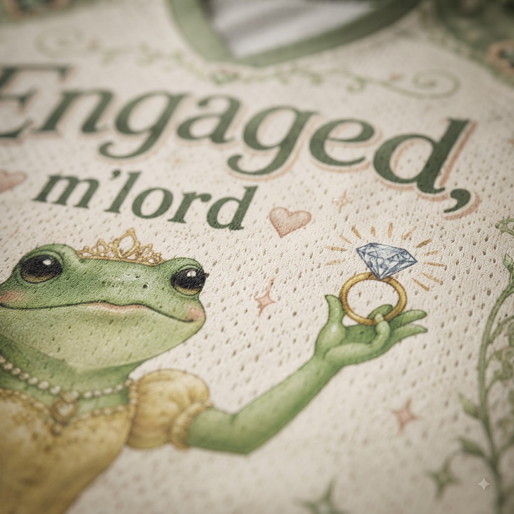 Close-up of a cream-colored mesh garment featuring a printed illustration of a green frog wearing a gold crown and holding a diamond ring, with the text 'Engaged, m'lord' in dark green script.