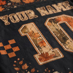 Close-up of a black mesh sports jersey with a personalized design, featuring a distressed tan 'YOUR NAME' text, a large number '10' filled with an autumn leaf camouflage pattern in orange, brown, and beige, and small orange leaf motifs near the bottom.