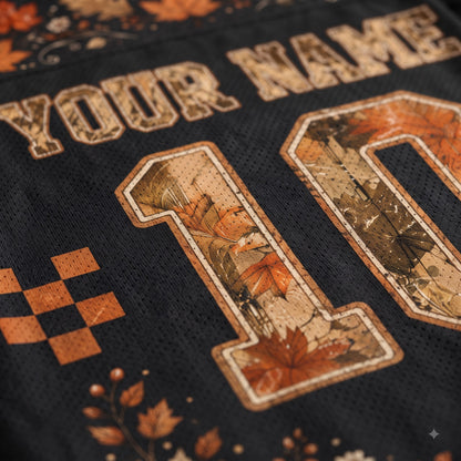 Close-up of a black mesh sports jersey with a personalized design, featuring a distressed tan 'YOUR NAME' text, a large number '10' filled with an autumn leaf camouflage pattern in orange, brown, and beige, and small orange leaf motifs near the bottom.