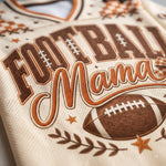 Close-up of a cream-colored athletic jersey with a 'Football Mama' graphic in brown and orange, featuring a football and star details.