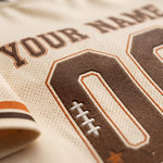 Close-up of a custom sports jersey featuring a cream-colored mesh fabric with brown and orange athletic lettering and numbers, including a football patch detail.