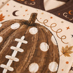 Close-up of a printed textile featuring a brown football-shaped pumpkin with white polka dots and white laces, on a cream background with autumn motifs like stars, a crescent moon, and leaves.