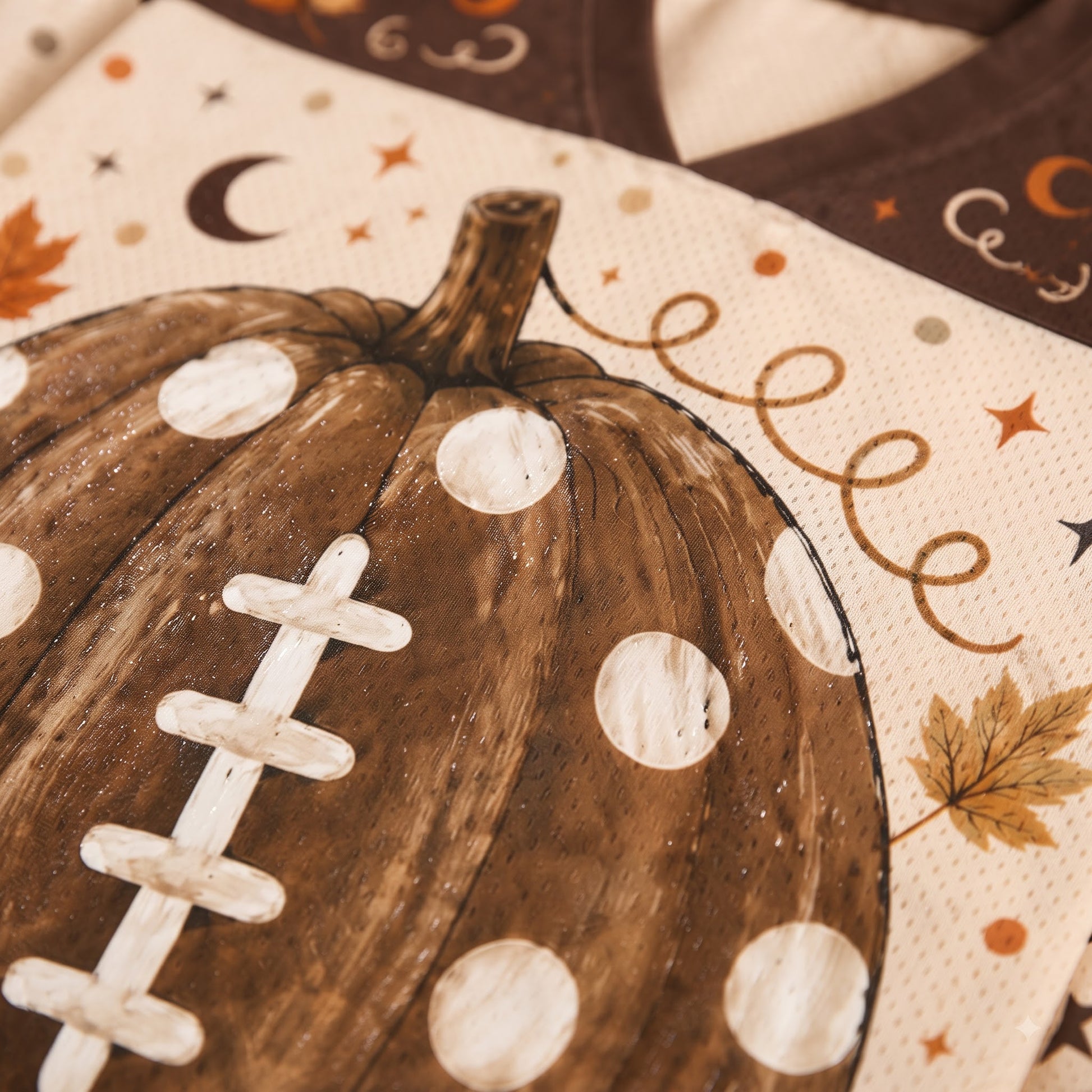 Close-up of a printed textile featuring a brown football-shaped pumpkin with white polka dots and white laces, on a cream background with autumn motifs like stars, a crescent moon, and leaves.