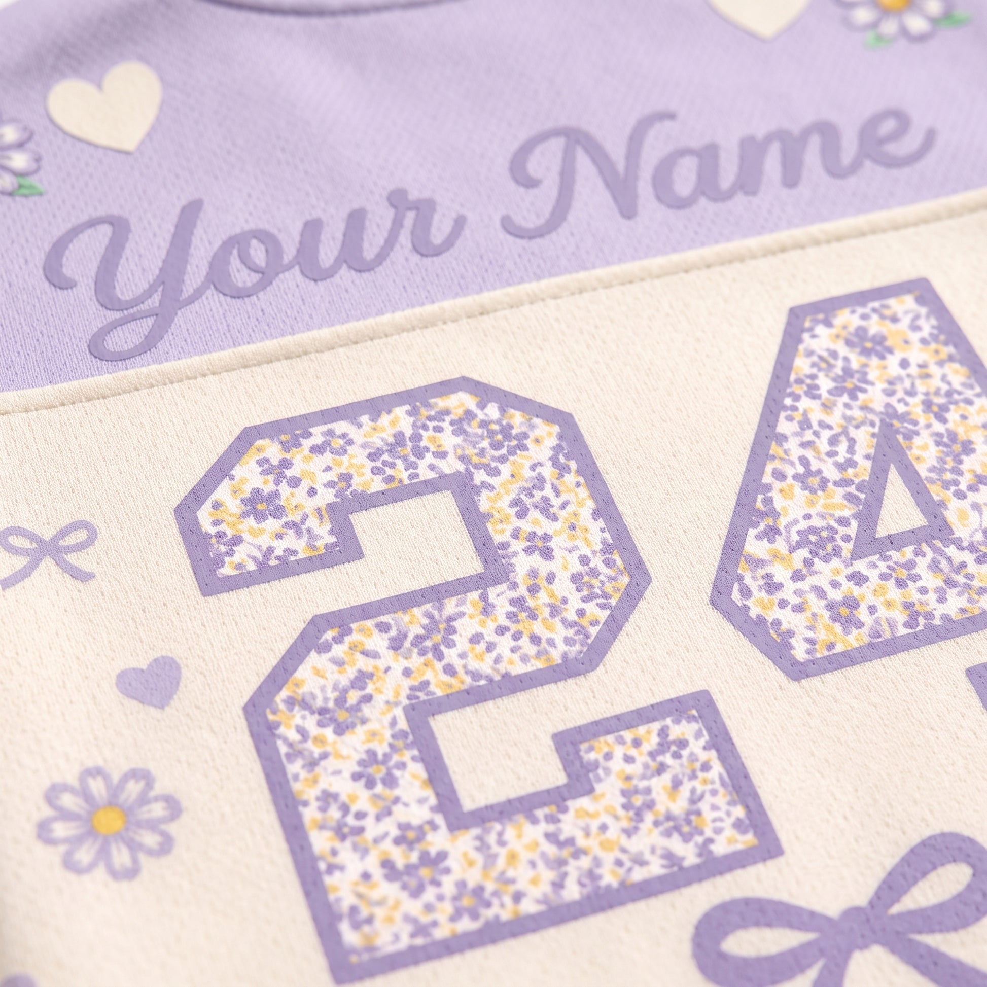 Close-up of a personalized sports jersey fabric with lavender purple text reading 'Your Name' and floral-patterned numbers on a cream background.