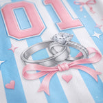 A close-up graphic design featuring two silver rings with a large clear marquise gemstone and smaller round stones, tied with a pink ribbon, on a white and light blue striped background with pink '01' numerals and small heart and star motifs.