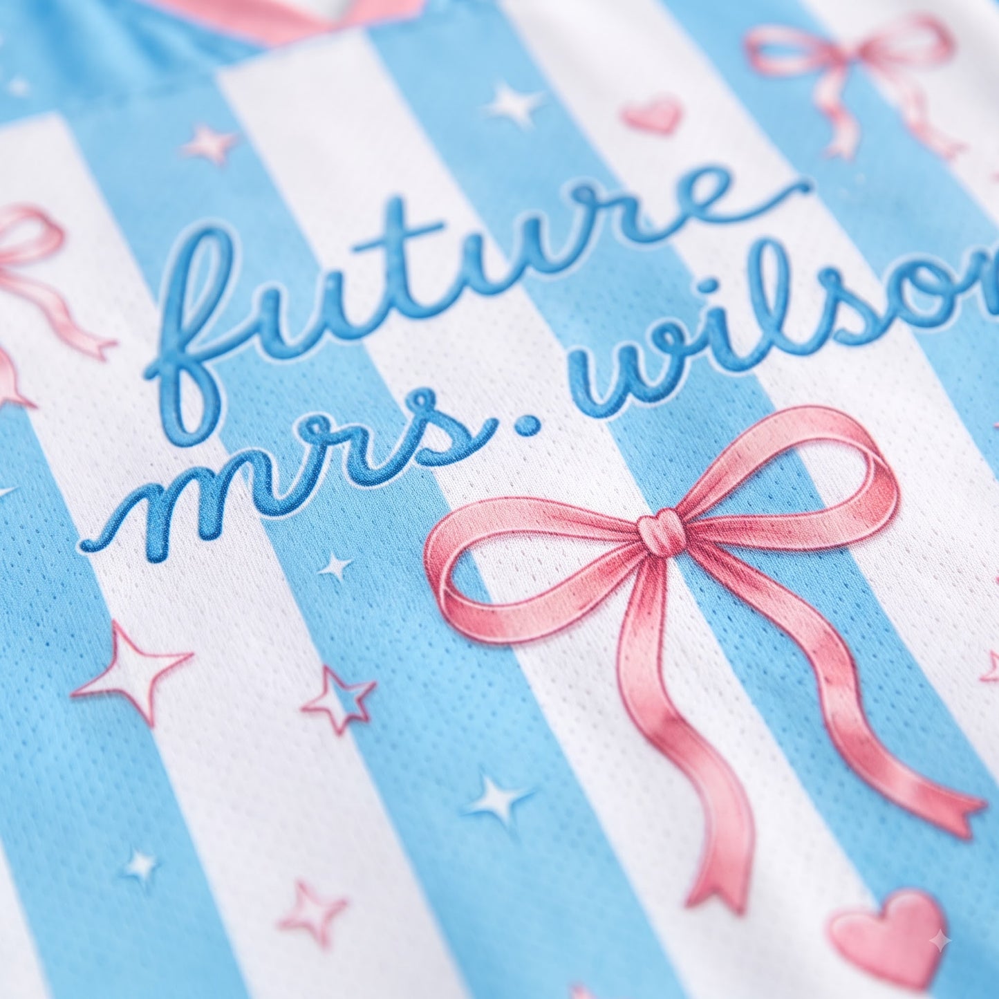 Close-up of a light blue and white striped garment featuring embroidered blue script reading 'future Mrs. Wilson' alongside pink bows, stars, and hearts.