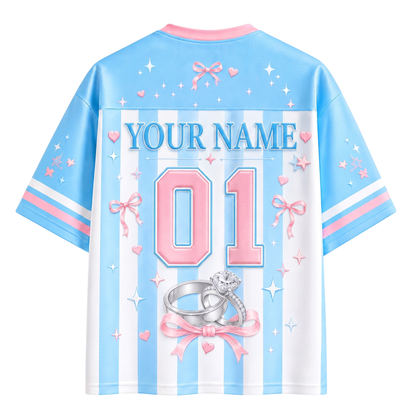 Customizable short-sleeve t-shirt shown from the back, featuring a light blue and white vertical stripe pattern with pink accents, including bows, hearts, stars, and a ring graphic. The back has a large customizable text area reading 'YOUR NAME' and a large number '01'.