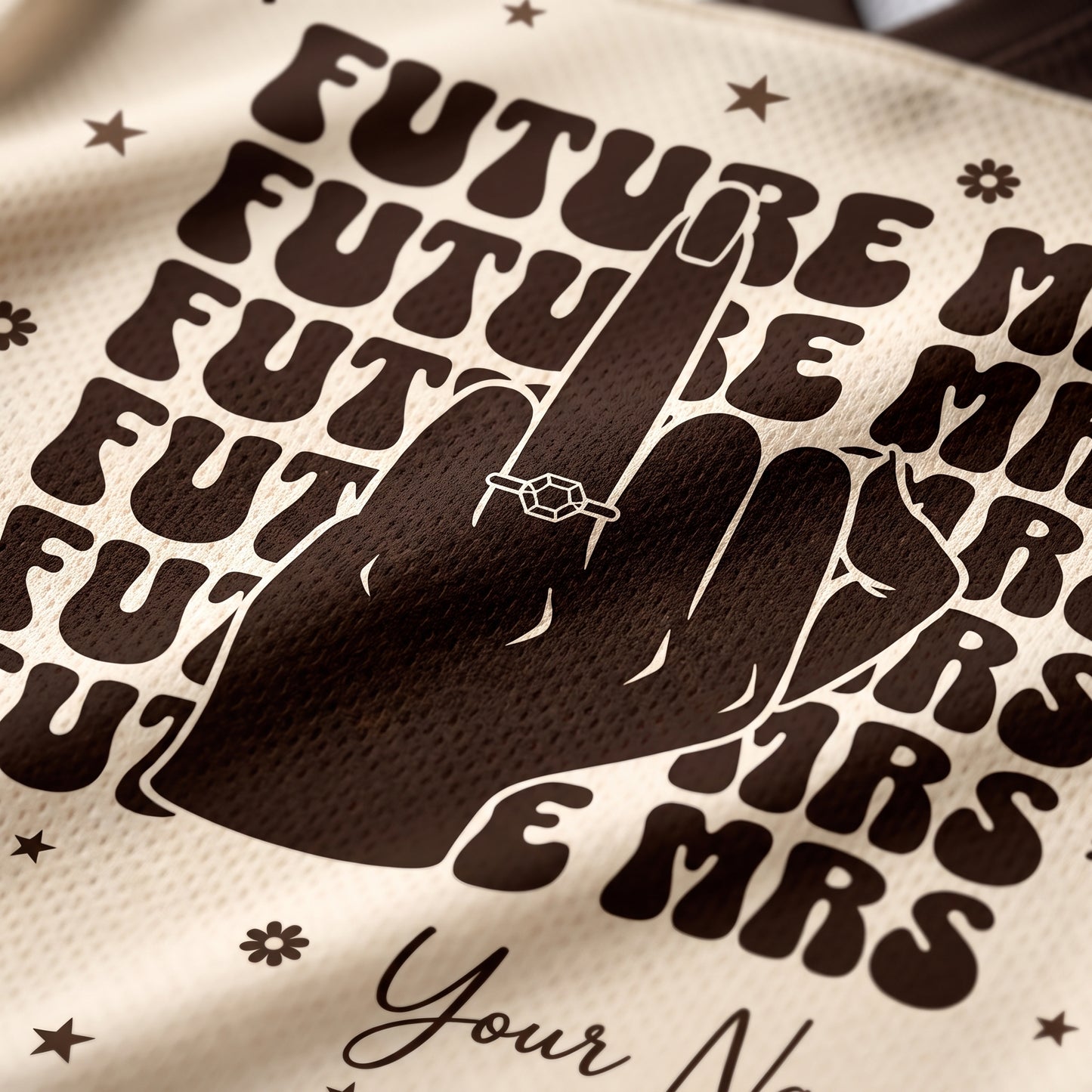 Close-up of a cream-colored mesh fabric with a dark brown graphic print featuring the word 'FUTURE MRS' repeated, a hand silhouette, stars, and partial script text reading 'Your N.