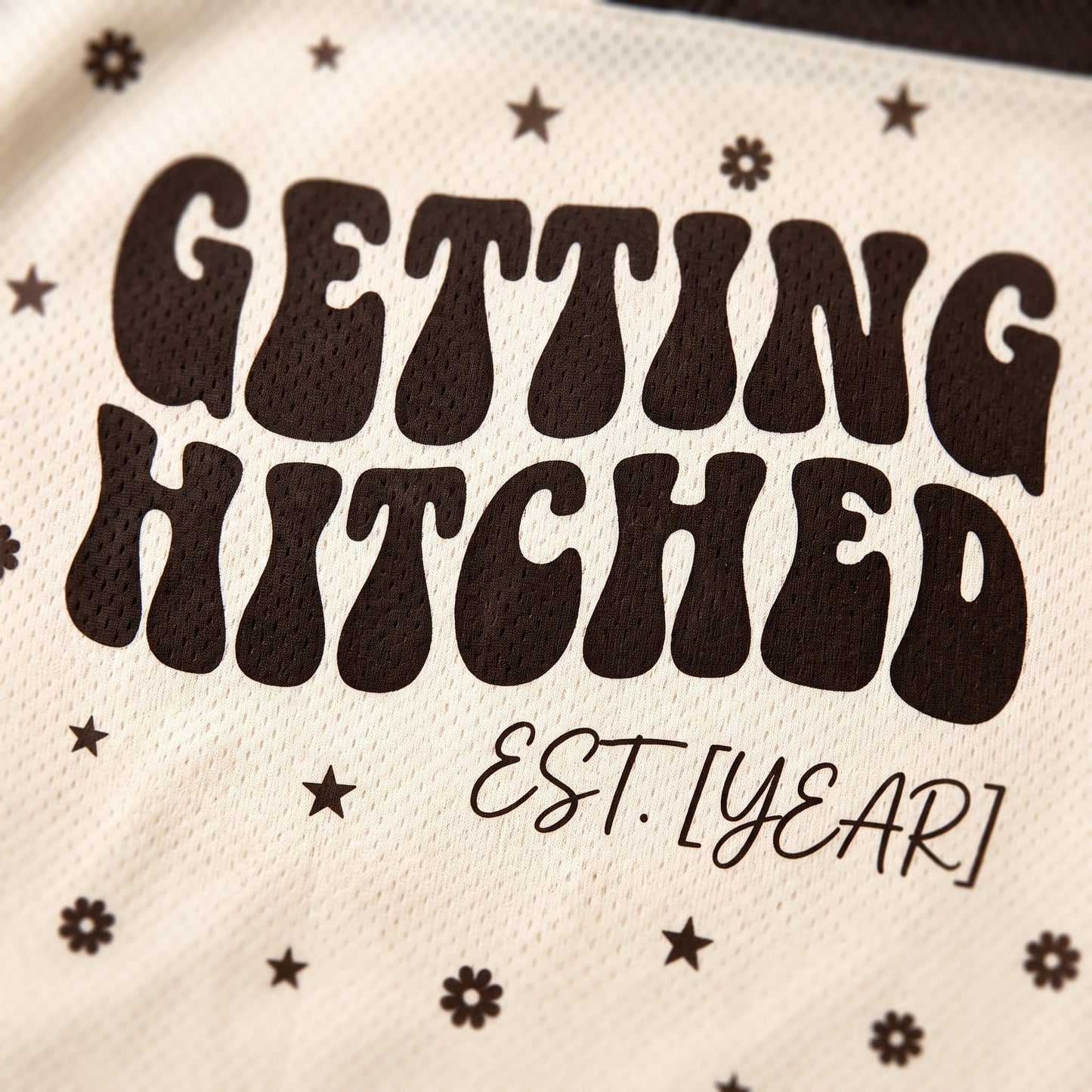 Close-up of a cream-colored mesh fabric with dark brown retro-style text reading 'GETTING HITCHED' and smaller script 'EST. [YEAR]', decorated with small star and flower motifs.