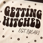 Close-up of a cream-colored mesh fabric with dark brown retro-style text reading 'GETTING HITCHED' and smaller script 'EST. [YEAR]', decorated with small star and flower motifs.